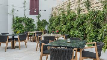 An outdoor seating area with tables and chairs, surrounded by lush greenery and a stone wall.