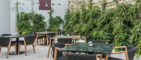 An outdoor seating area with tables and chairs, surrounded by lush greenery and a stone wall.
