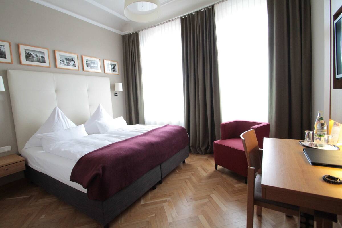 Une chambre d’hôtel avec un grand lit, un bureau avec une chaise, une lampe et une fenêtre avec des rideaux.