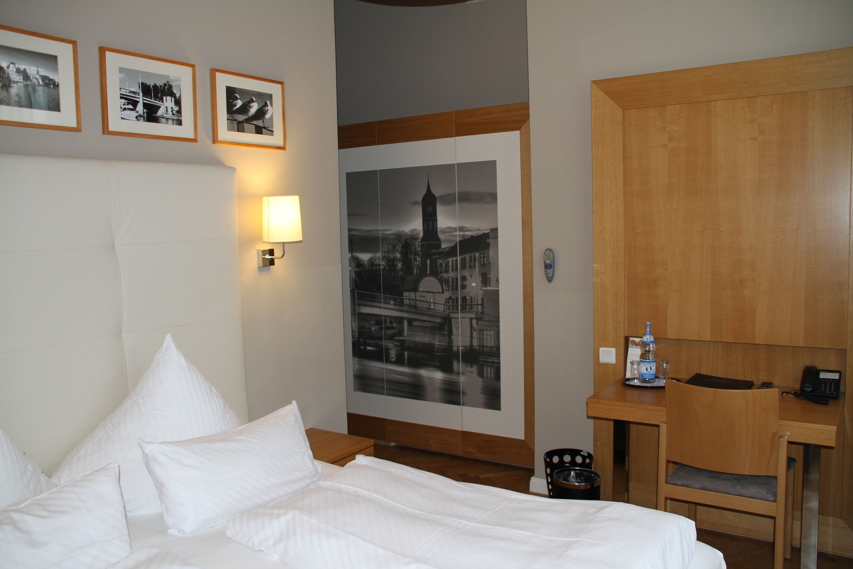A hotel room with a bed, a desk, a chair, a lamp, a framed picture, and a water bottle.