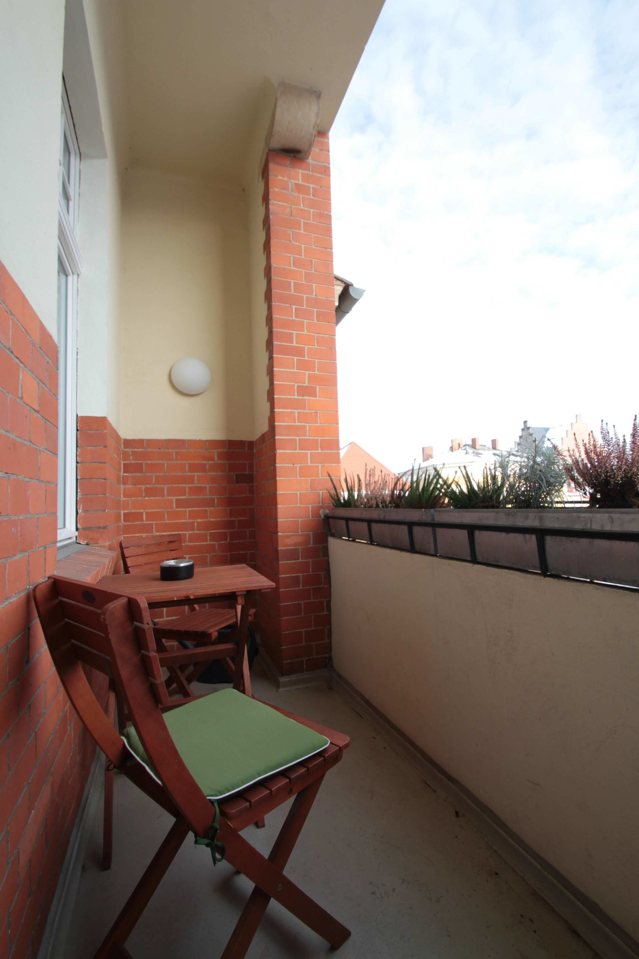 Un pequeño balcón con una mesa y sillas, un muro de ladrillo y vistas a edificios al fondo.