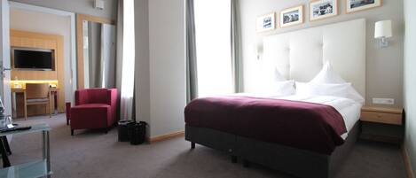 Una habitación de hotel con una cama grande, un sillón rojo, un escritorio con televisor y un ventanal con cortinas.