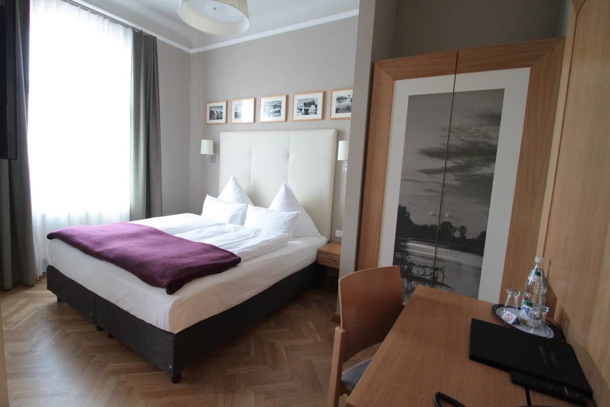Une chambre d’hôtel avec un lit, un bureau et une chaise. Les murs sont ornés de tableaux encadrés et il y a une fenêtre avec des rideaux.