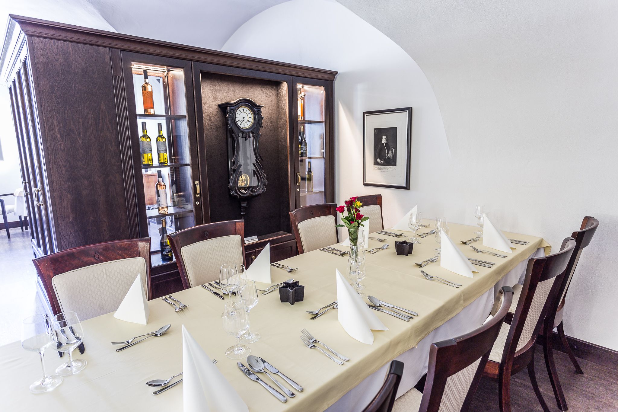 Uma sala de jantar com uma mesa comprida posta para refeição, cadeiras de madeira, um relógio e um quadro na parede.