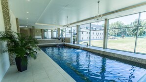 An indoor swimming pool with clear blue water, a staircase leading to a deck area, and large windows offering a view of the outdoors. - Chateau Appony (Oponice)
