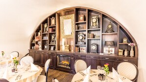 A dining area with round tables set for two, each with white tablecloths and silverware. There is a fireplace in the background, and the walls are lined with shelves holding various clocks and decorative items. - Chateau Appony (Oponice)