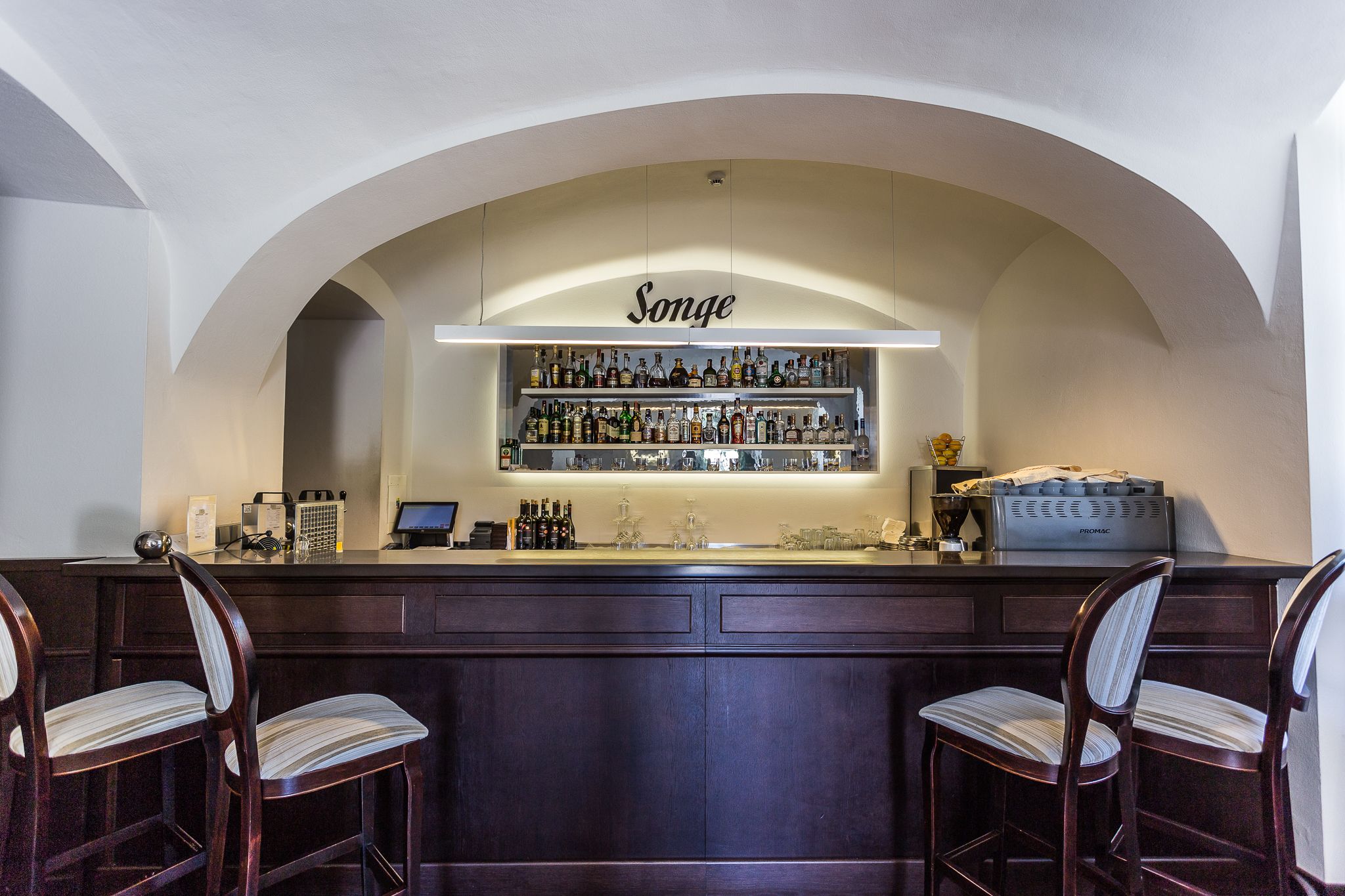 A bar area with a 'Songe' sign above shelves of bottles, a coffee machine, and two bar stools.
