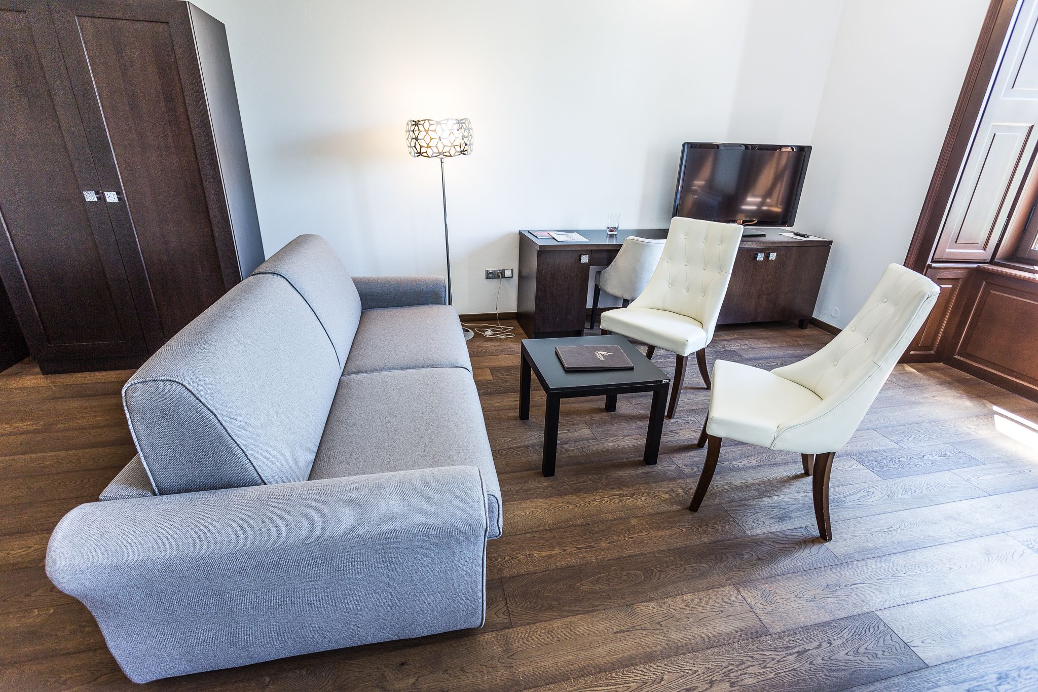 A modern living room with a grey sofa, two white armchairs, a small table, a floor lamp, and a TV.