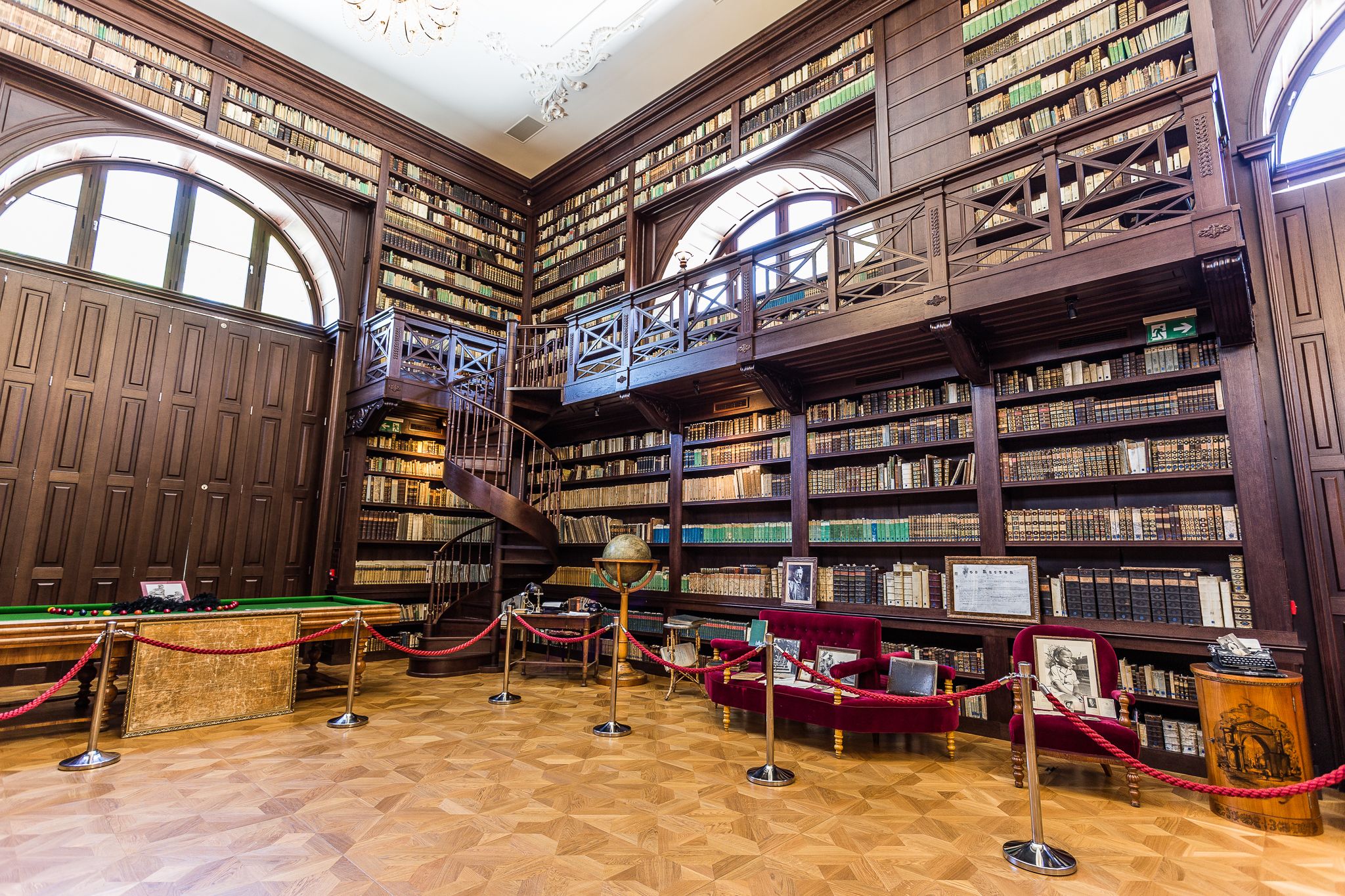 A large, ornate library with multiple levels of bookshelves, a grand staircase, and a billiards table.
