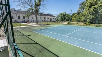 A tennis court with a green fence, a building in the background, and trees around.