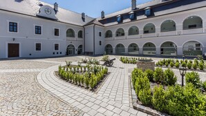 A courtyard with a cobblestone path, a cannon, and a building with arched windows and a clock. - Chateau Appony (Oponice)