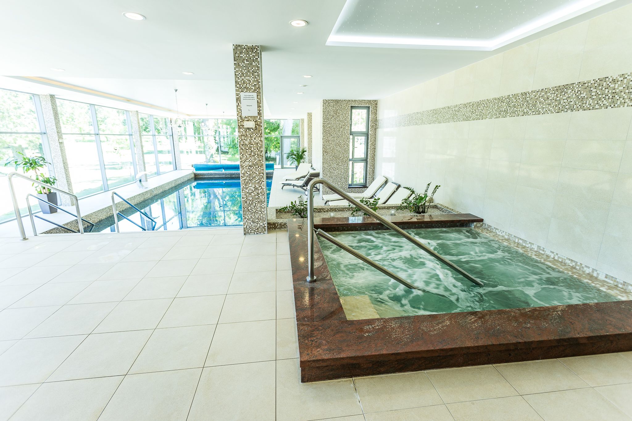A modern indoor pool area with a clear view of the pool, a jacuzzi, and a sun terrace with lounge chairs.