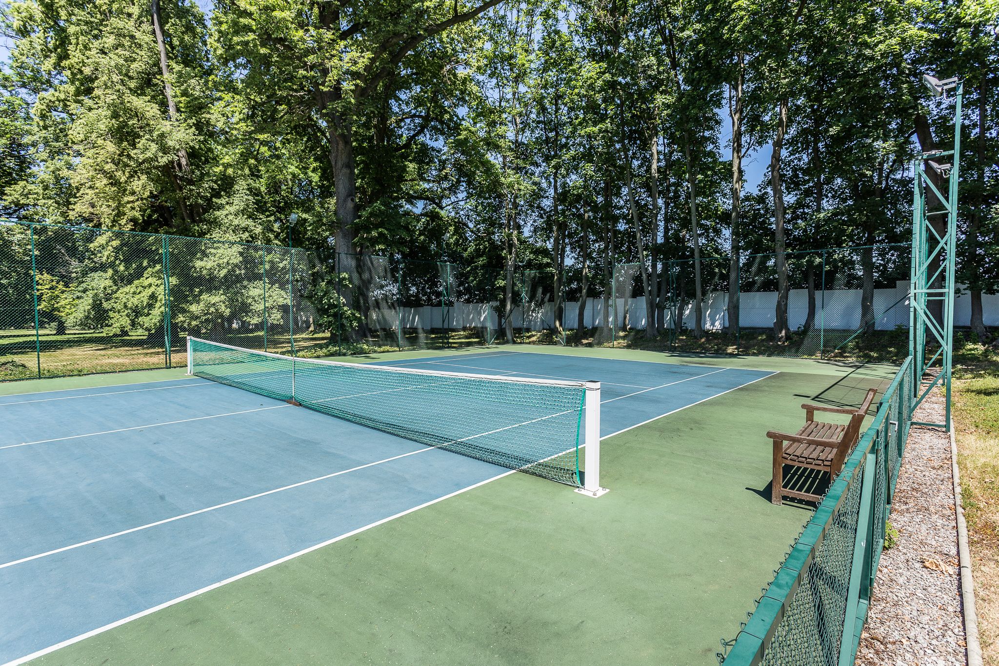 An outdoor tennis court with a net, surrounded by a green fence and trees.