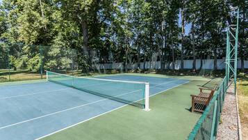 An outdoor tennis court with a net, surrounded by a green fence and trees.