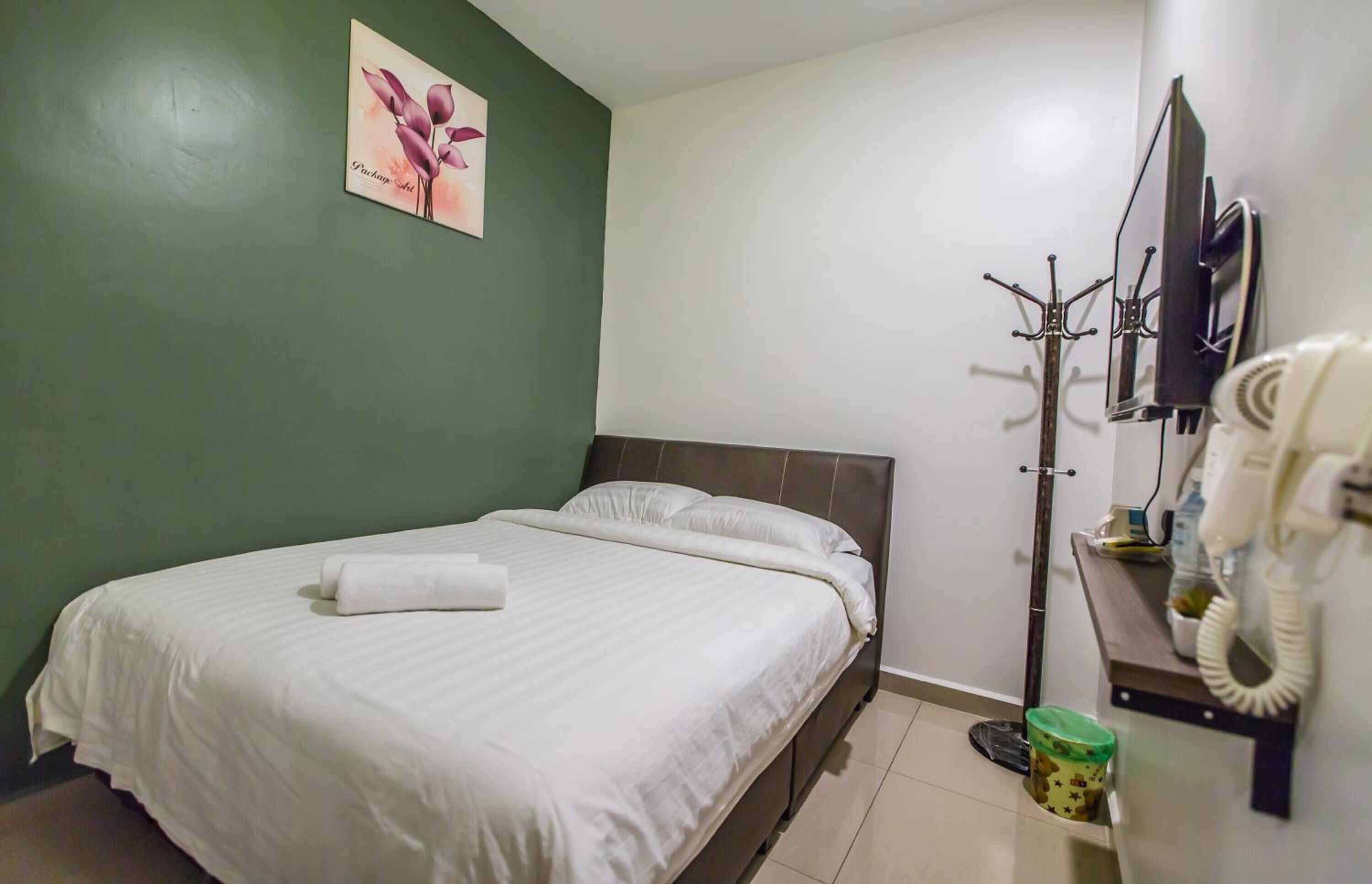 A neatly made bed with a white comforter and pillows, a green wall with a floral painting, a coat rack with a coat and a hat, and a telephone on a desk.