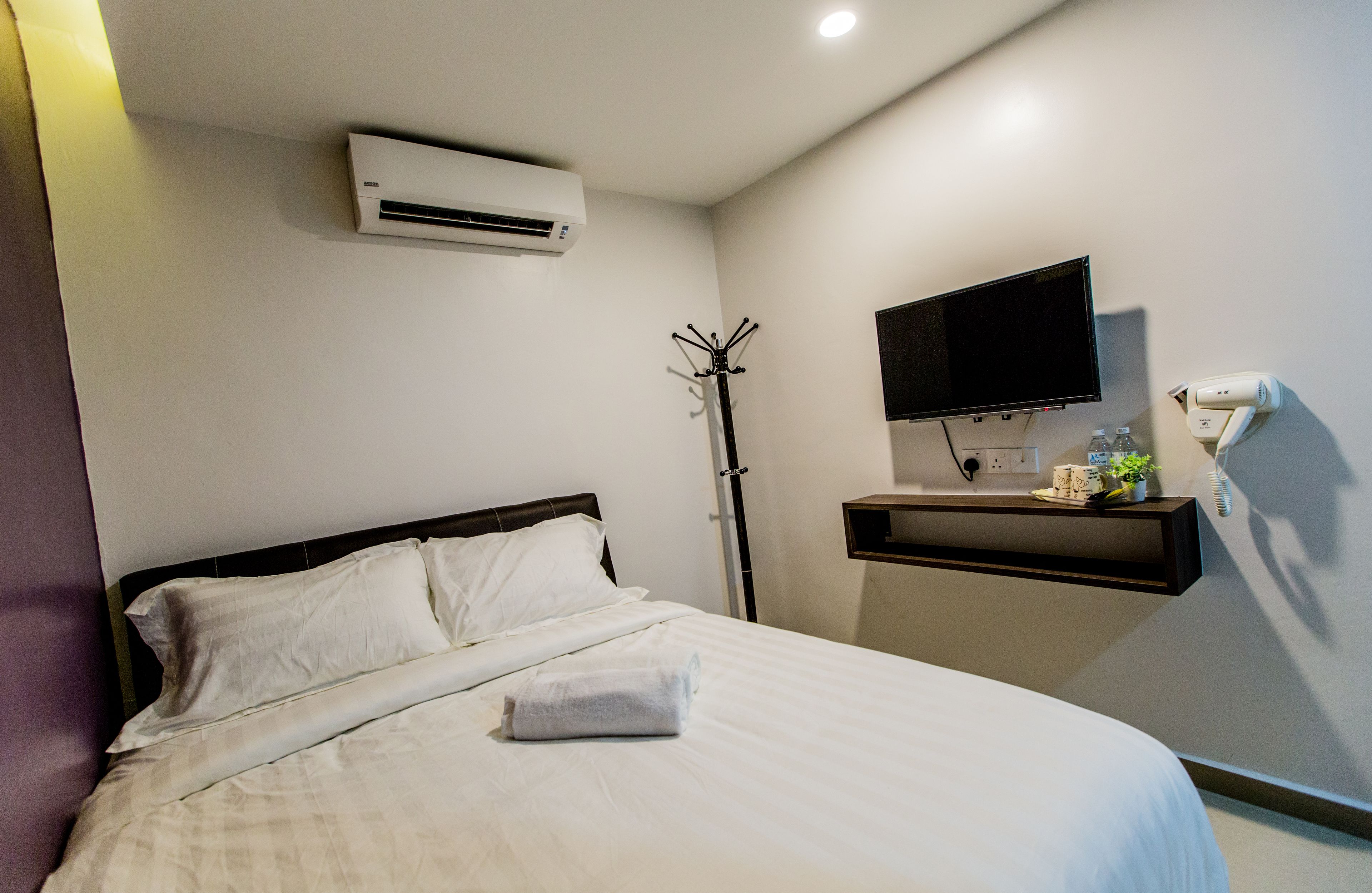 A hotel room with a bed, an air conditioning unit, a mounted TV, a coat rack, and a bedside table with a plant and a water bottle.