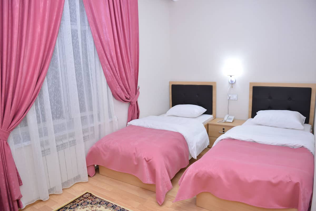 A room with two beds, each with a white headboard and pink bedding. There is a nightstand with a phone and a lamp on each bed. The room has a wooden floor and a window with pink curtains.