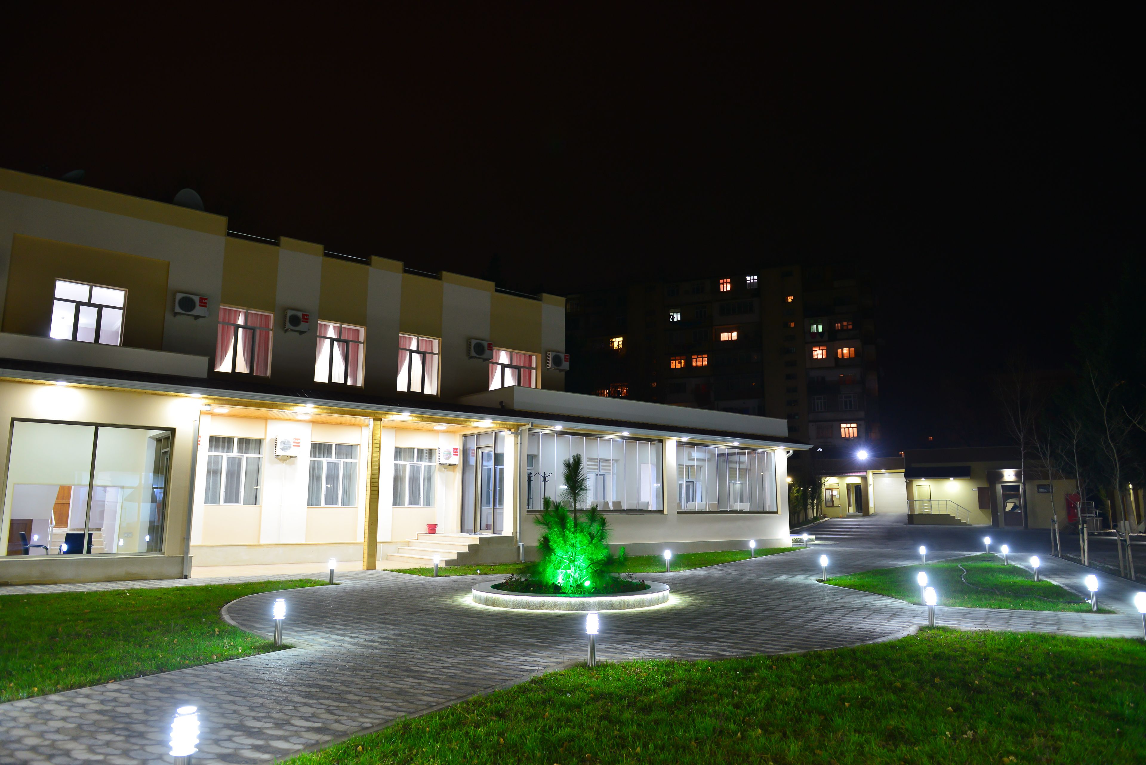 A building with multiple windows and air conditioning units, a paved walkway, and a circular garden with lights.