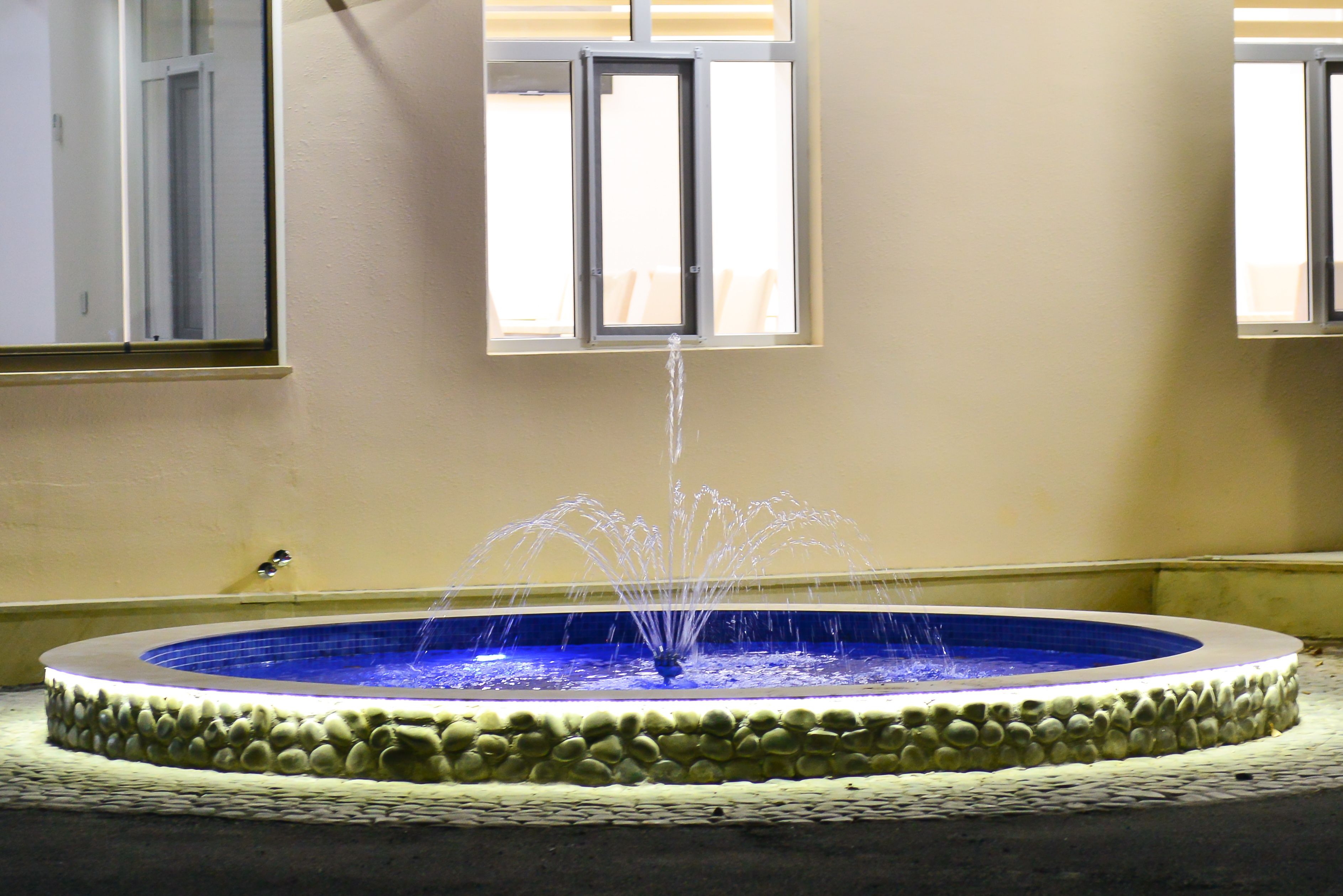 A circular fountain with water jets, surrounded by a stone border, and a window above it.