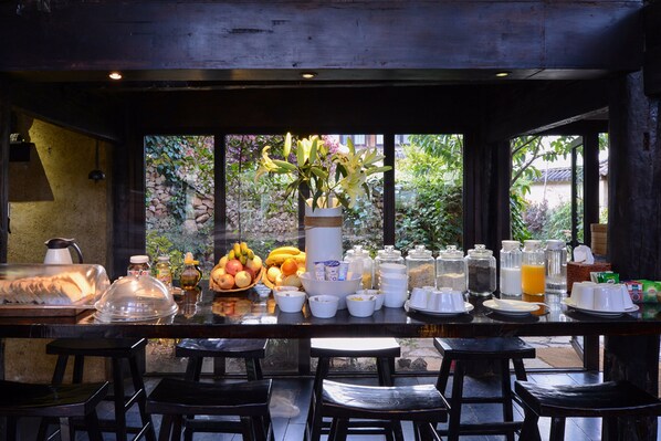 A breakfast bar with a variety of food items, including fruits, bread, and beverages, and a view of greenery outside.