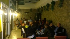 Un grupo de personas sentadas en mesas en un espacio exterior para comer, con paredes de piedra y macetas con plantas.