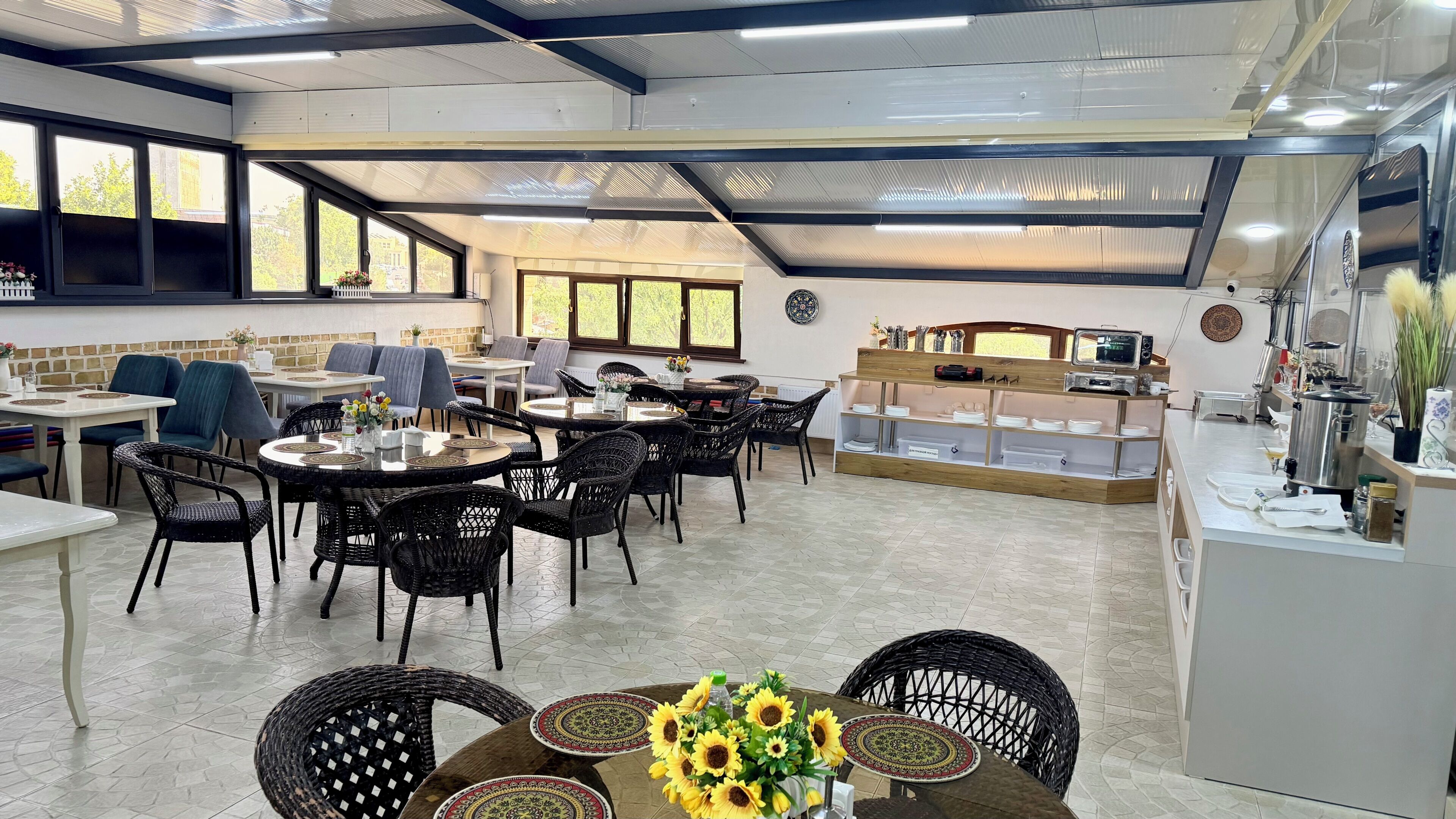 A spacious indoor dining area with multiple tables and chairs, a kitchen counter, and a view of the outdoors through large windows.