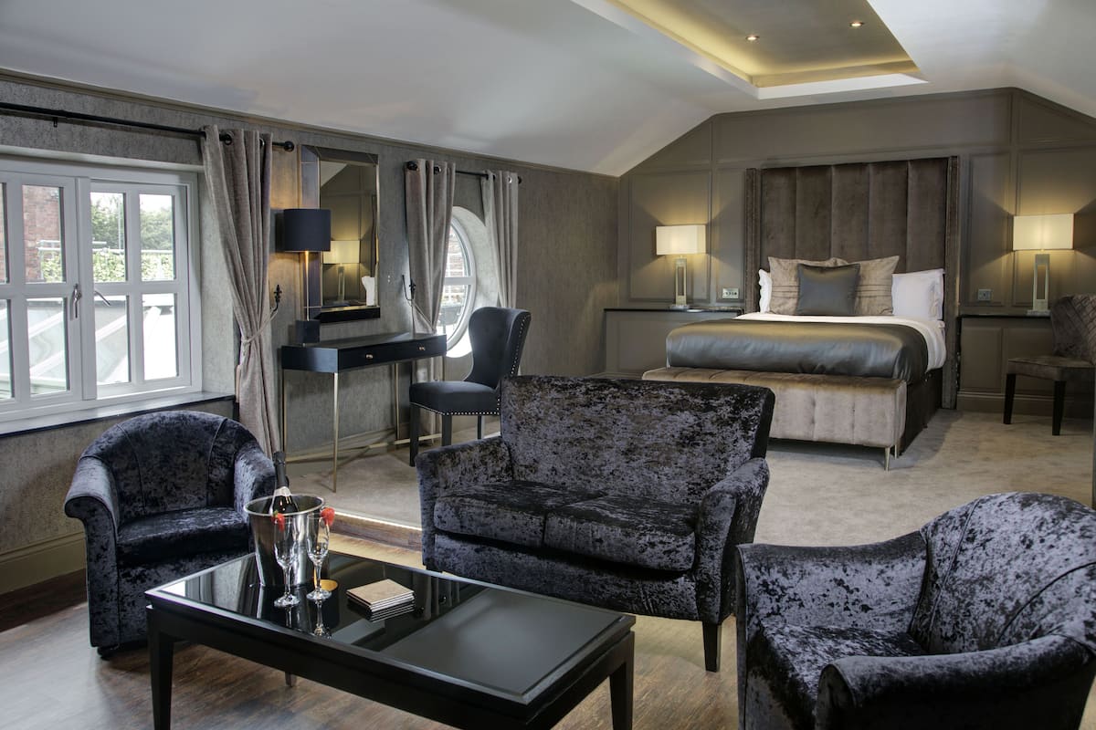 A hotel room with a large bed, two armchairs, a desk, and a glass table with a bottle and glasses.
