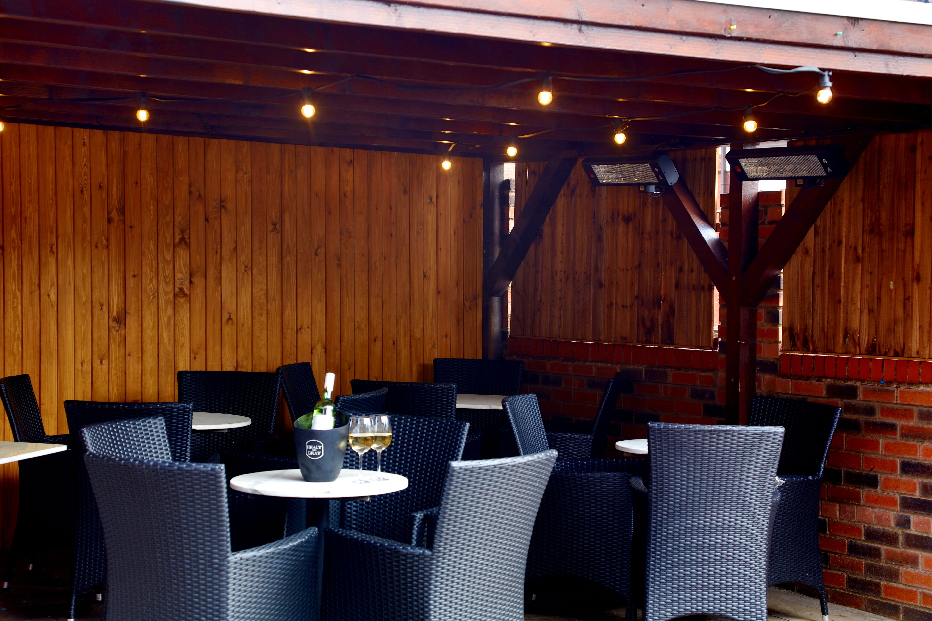 Un espace extérieur aménagé avec des chaises et des tables en osier, un mur en briques et des poutres en bois.