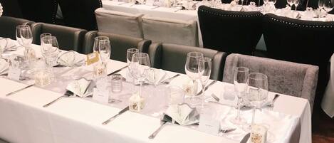 Une salle à manger élégante, avec de longues tables dressées pour accueillir de nombreux convives, nappes blanches, couverts disposés avec soin et verres à vin.