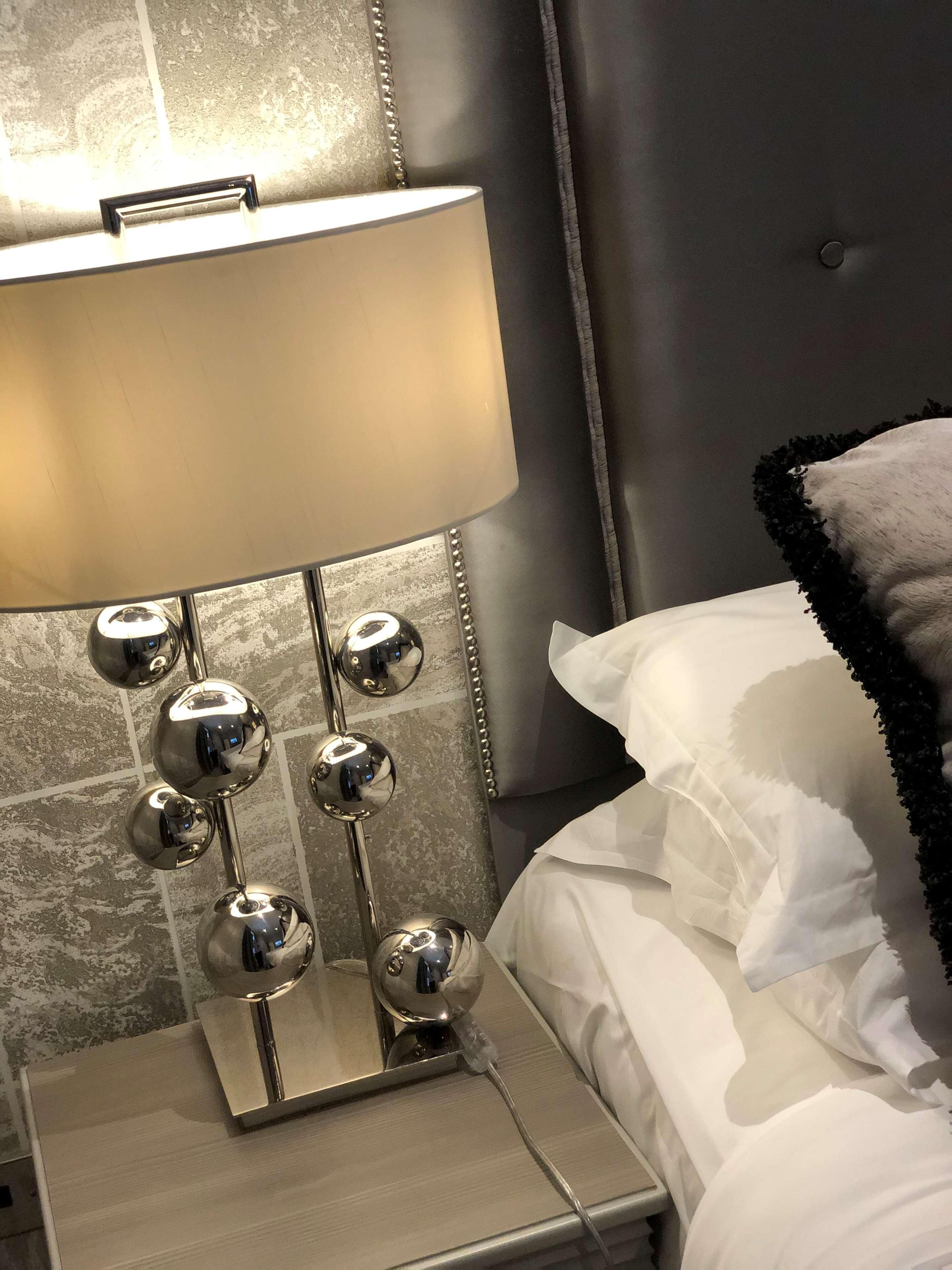 A bed with white linens, a grey headboard, and a bedside lamp with multiple spherical mirrors.