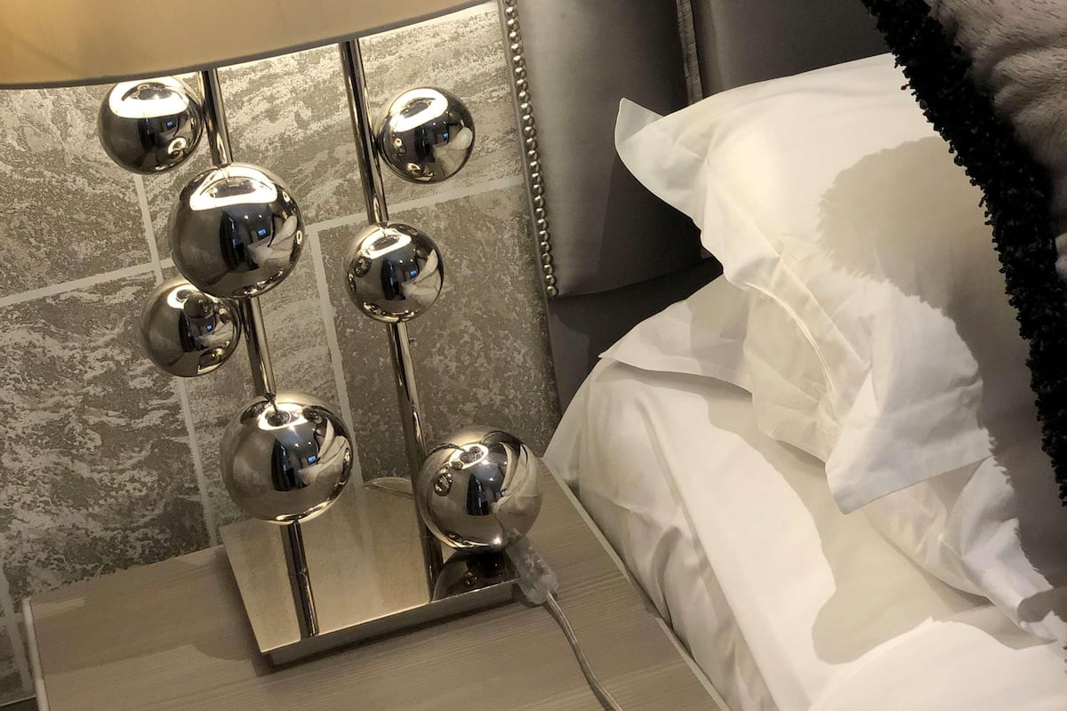 A bed with white linens, a grey headboard, and a bedside lamp with multiple spherical mirrors.