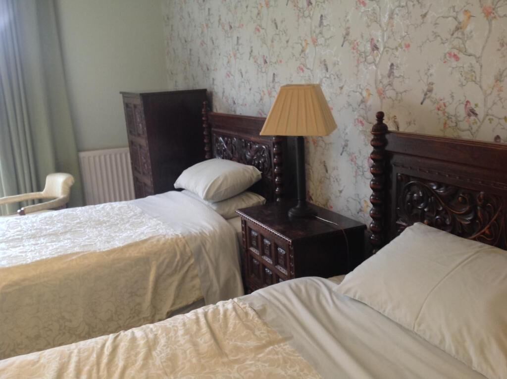 Quarto de hotel com duas camas, painel de cabeceira em madeira, mesa de cabeceira com abajur e papel de parede floral.