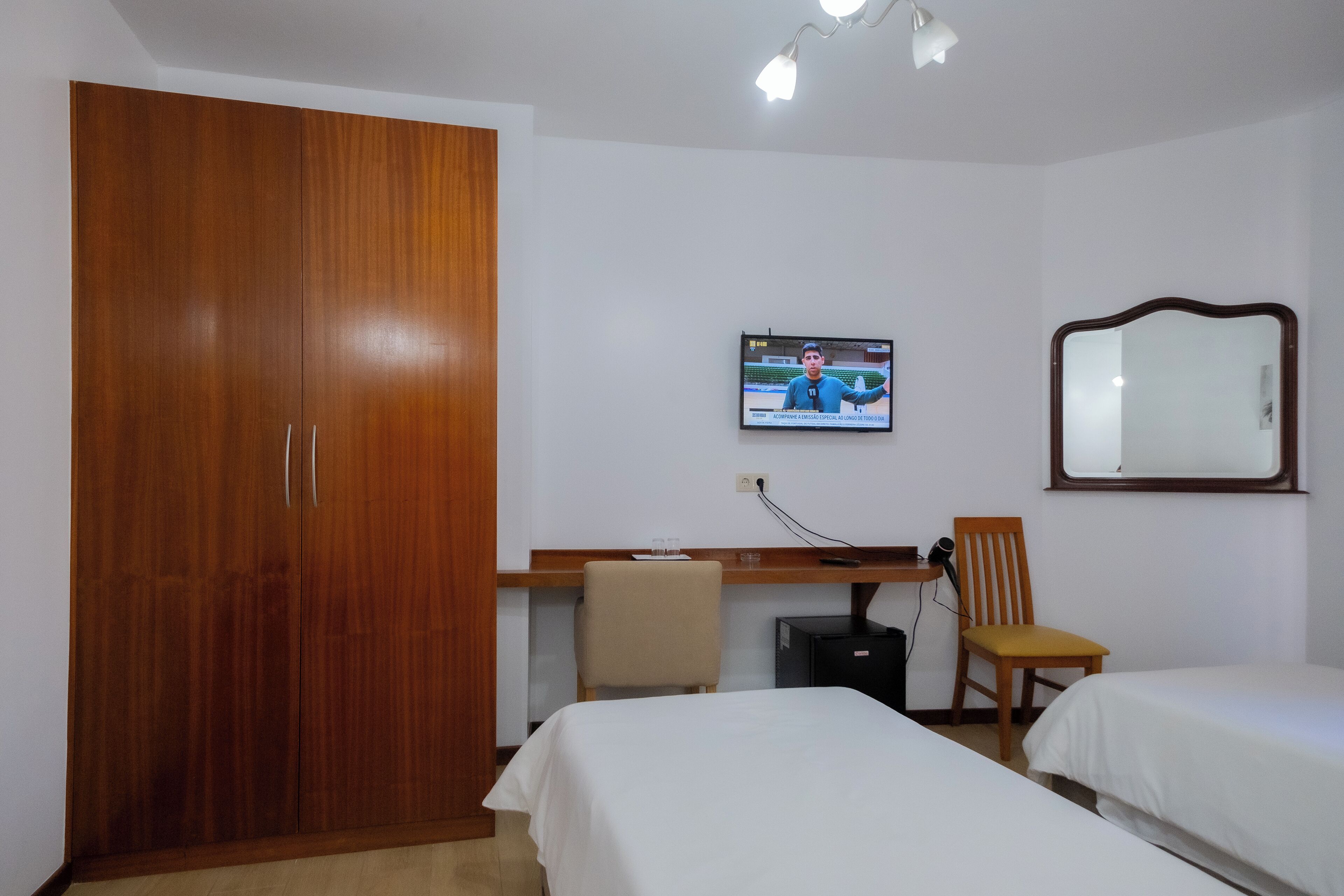 A hotel room with a wooden wardrobe, a desk with a chair, a television, and two beds.