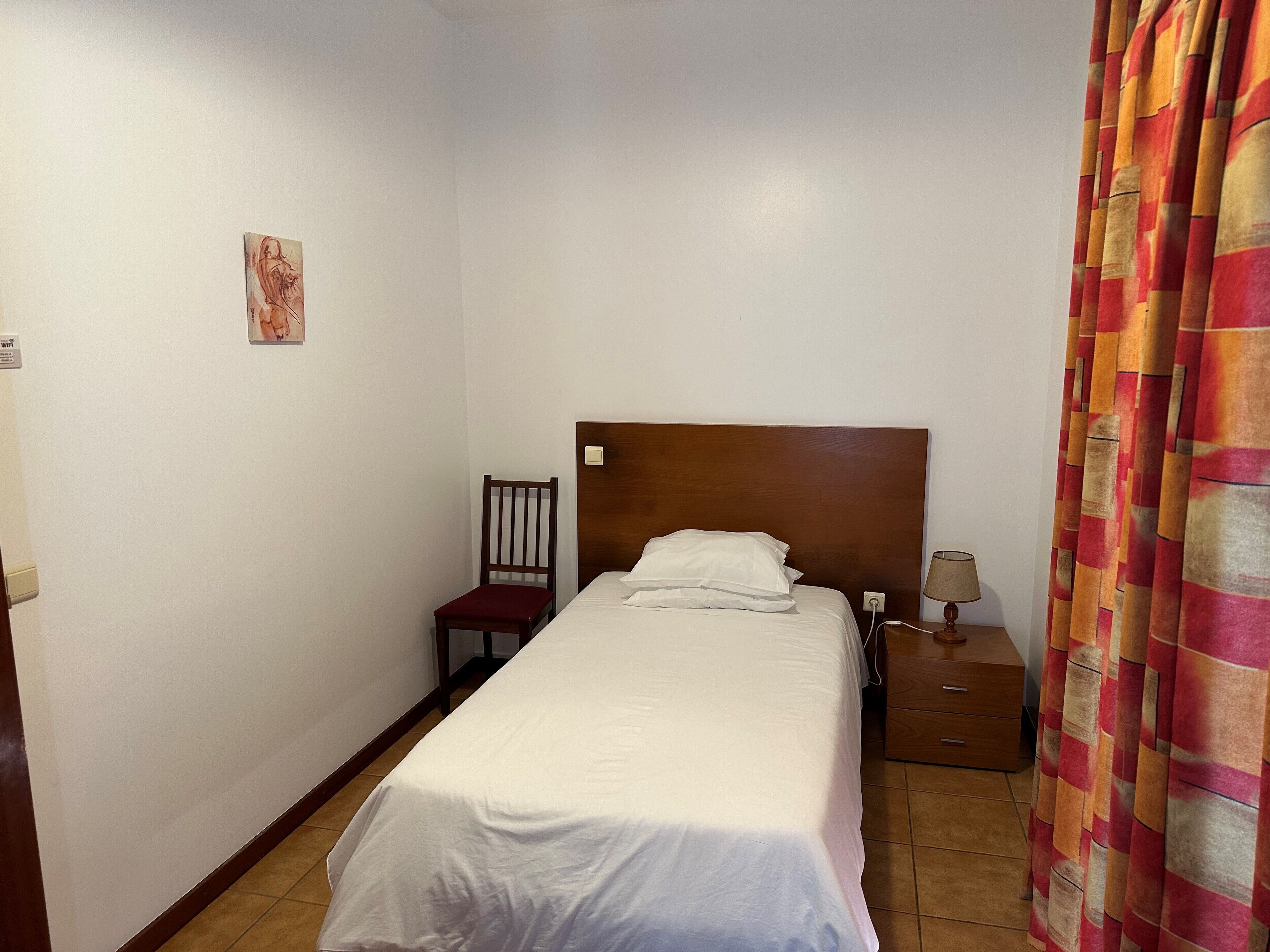 A single bed with a wooden headboard, a chair, a nightstand with a lamp, and a curtain.
