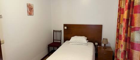 A single bed with a wooden headboard, a chair, a nightstand with a lamp, and a curtain.