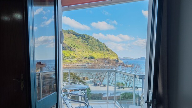 Puerta de vidrio abierta que da a un balcón con vistas a un paisaje costero, incluyendo una colina y el mar.