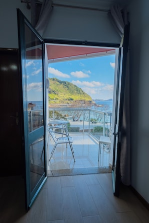 An open glass door leading to a balcony with a view of a coastal landscape, including a hill and the sea. - Residencial Família (Machico)