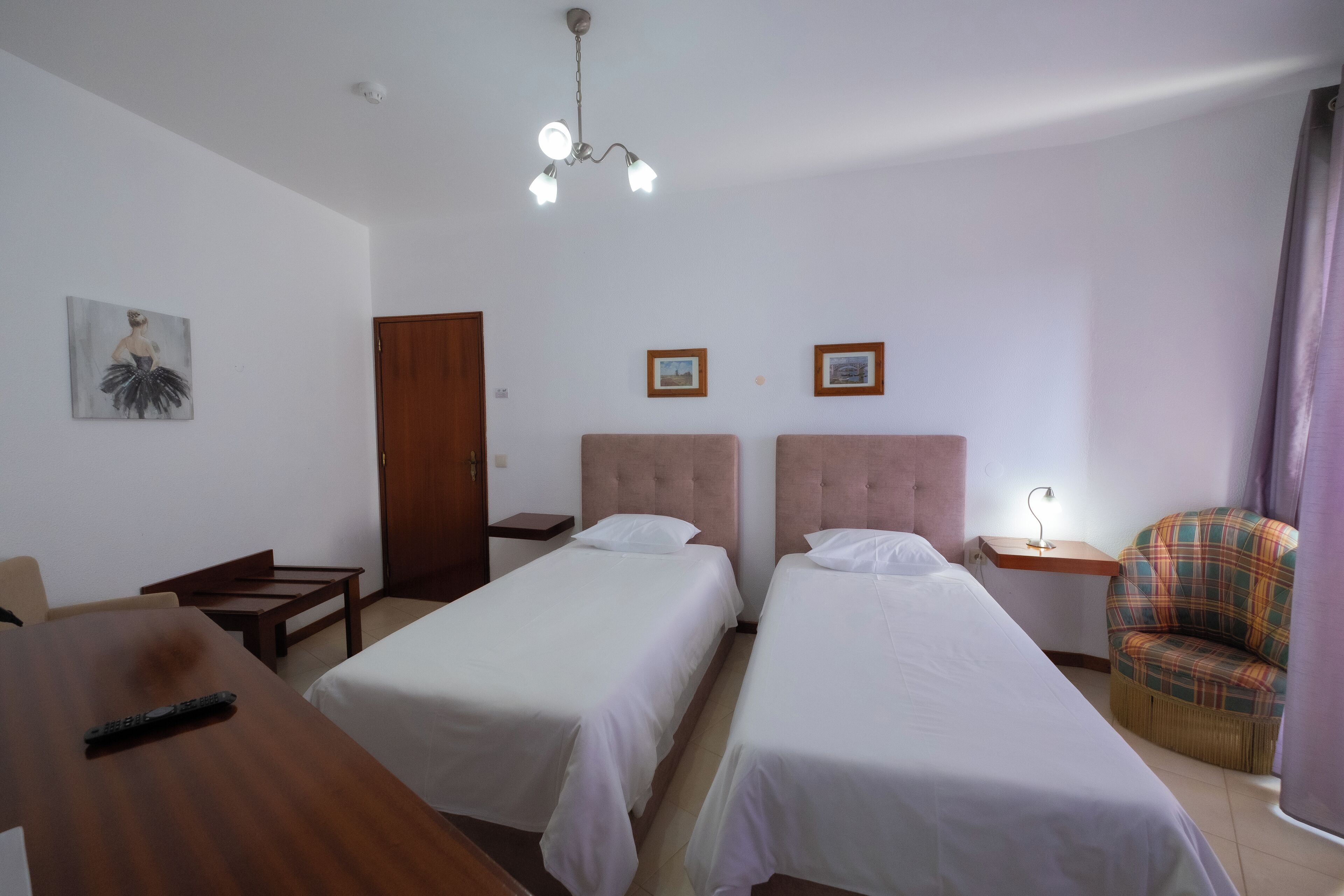 A hotel room with two single beds, a wooden desk, a chair, a small table, and a ceiling light fixture.