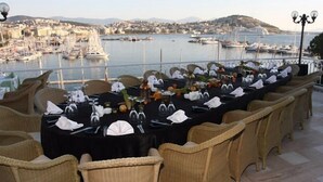 A dining table set for a meal with wicker chairs, overlooking a marina with boats and a coastal town in the background. - Kismet Hotel (Kusadasi)