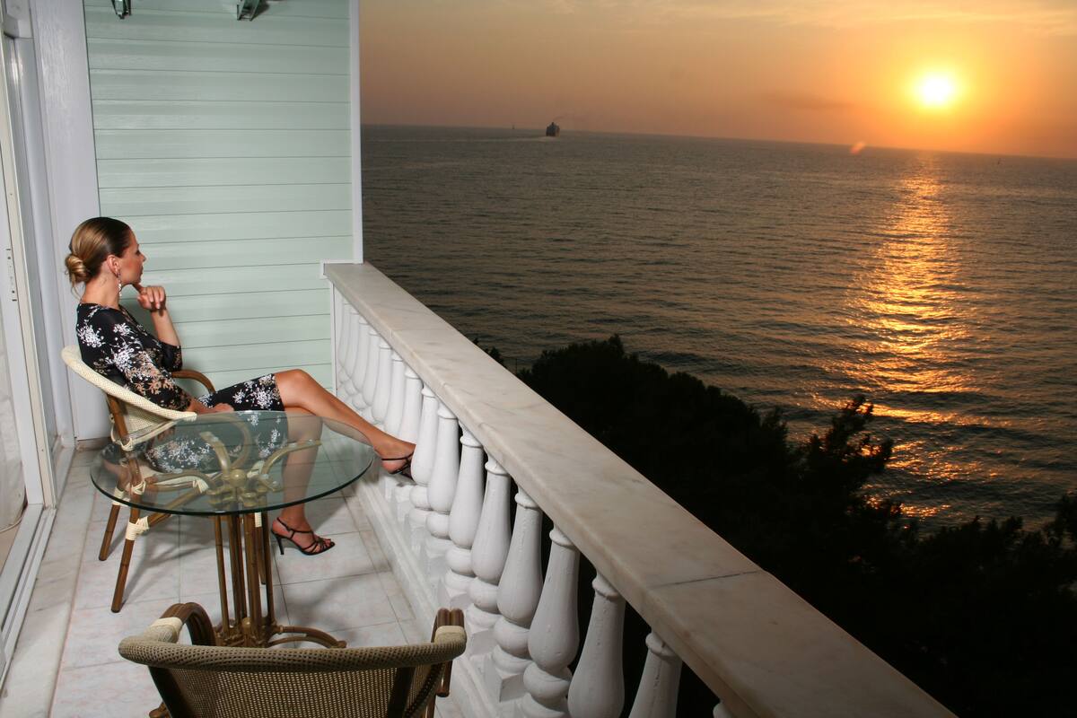 A balcony with a glass table and wicker chairs, overlooking the sea with a sunset view.