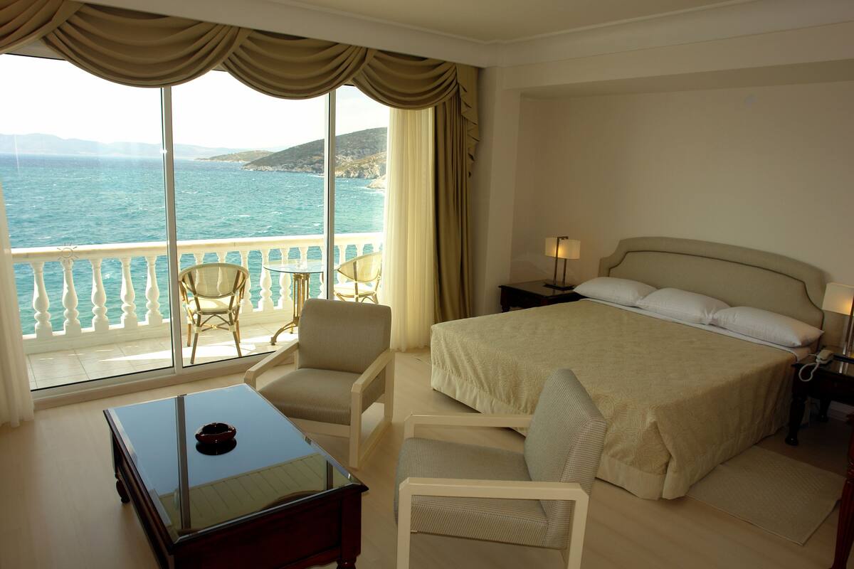 A bedroom with a large bed, two chairs, a desk, and a view of the sea through large windows.