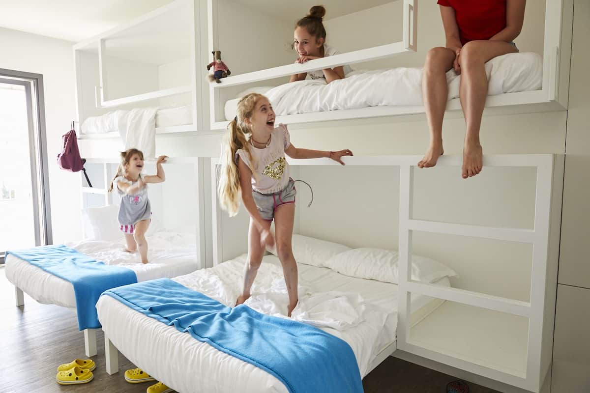 Een slaapkamer met stapelbedden, waar twee kinderen spelen en een vrouw op het bovenste bed zit.