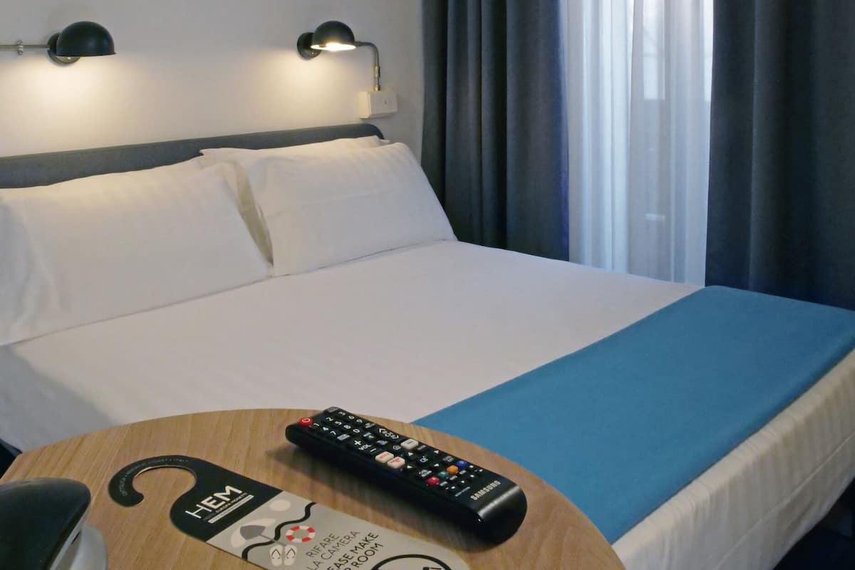 Een hotelkamer met een bed, twee bedlampen, een afstandsbediening en een bordje met informatie over faciliteiten zoals een minibar en een verzoek om de kamer op te maken.