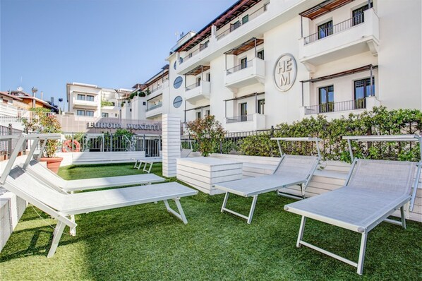 Une cour avec des chaises longues blanches et une piscine, entourée d’un bâtiment blanc doté de balcons et d’une enseigne sur laquelle on peut lire « EUROPA MONETTI ».