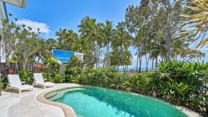 A pool area with lounge chairs, surrounded by tropical plants and palm trees, with a sign for Cairns.