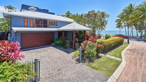 A modern two-story house with a garage, surrounded by well-maintained gardens and palm trees, near a beach.