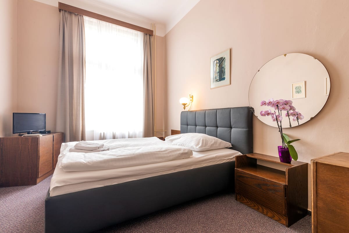 Ein Hotelzimmer mit Bett, Fernseher, Nachttisch mit Lampe, Spiegel und Blumen in einer Vase.