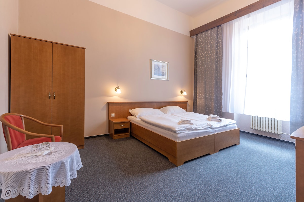 Ein Hotelzimmer mit einem Bett, einem Holzschrank, einem Nachttisch, einem Tisch mit weißen Spitzenstoffen und einem Fenster mit Vorhängen.