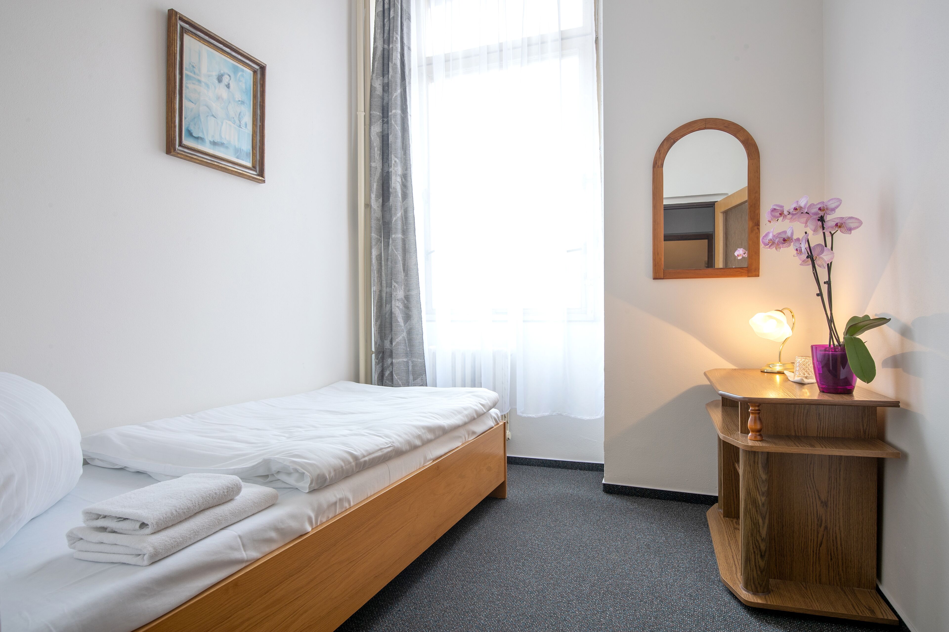 En pænt redt seng med hvide sengetøj, et trænatbord med en lampe og en lilla vase, og et spejl på væggen.