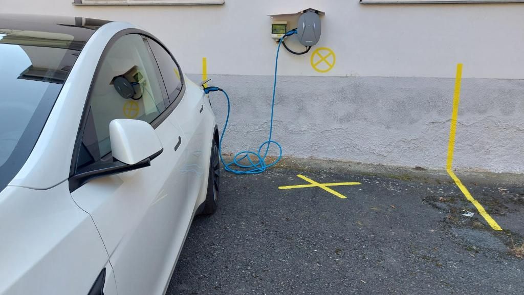 A white car parked next to a building with a charging station and yellow markings on the ground.